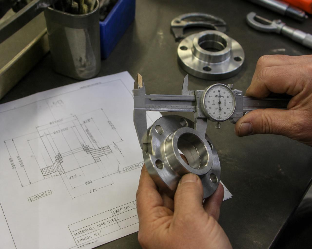 Machinist measuring a finished machined steel part with a dial caliper beside its drawing